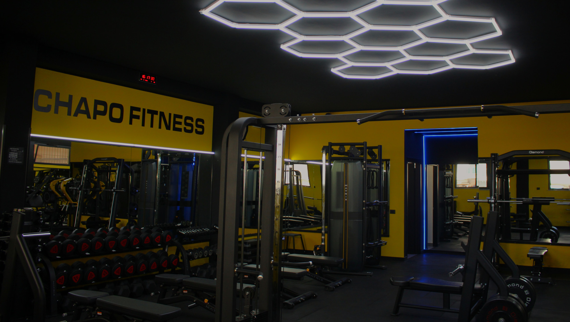Vista d'insieme della sala pesi di Chapo Fitness al Villaggio Santa Rosalia con insegna CHAPO FITNESS e illuminazione esagonale a LED